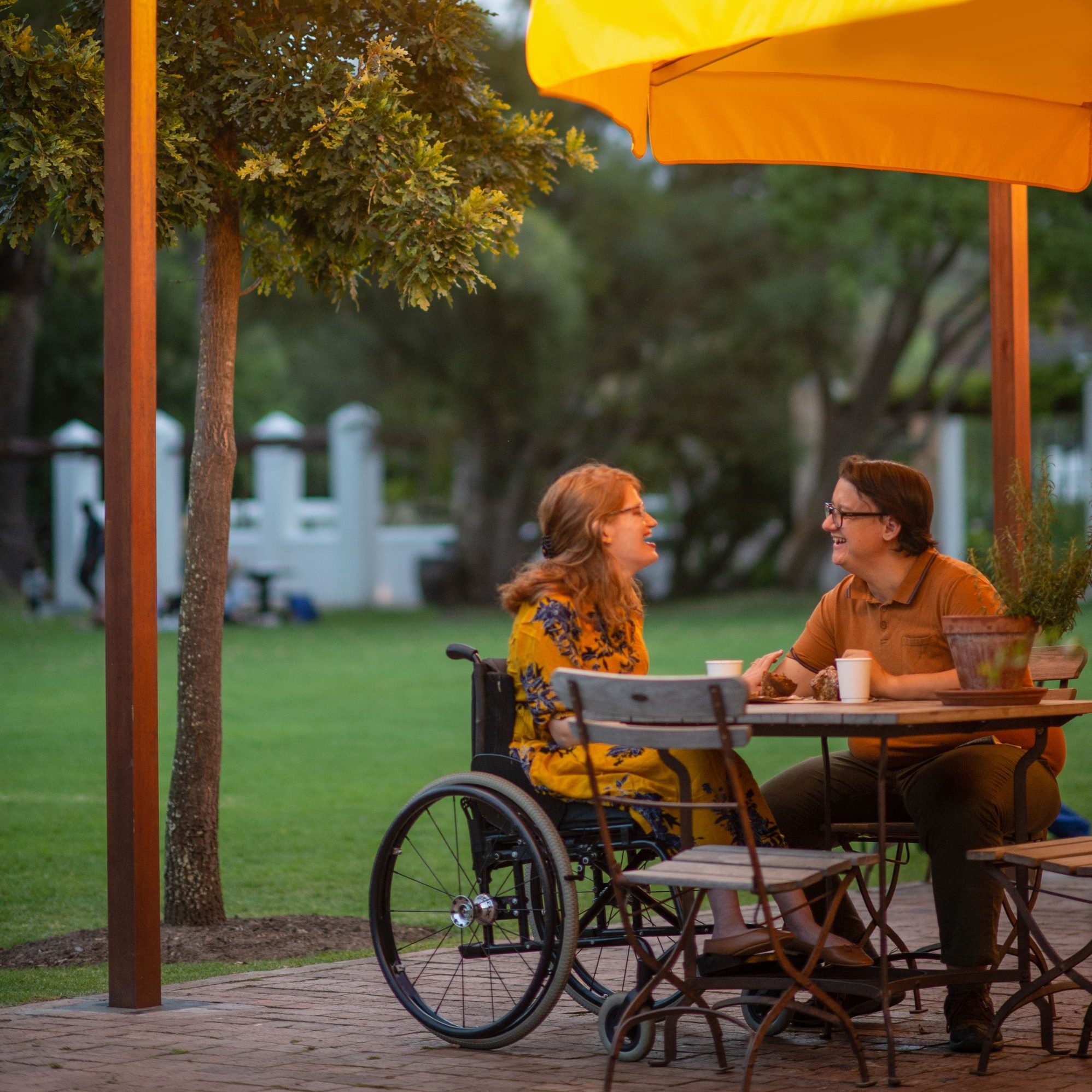 A Caucasian male and a disabled female with cerebral palsy in a wheelchair sitting outside at a restaurant cafe having take away coffees and muffins sitting under an umbrella and string lights at dusk.